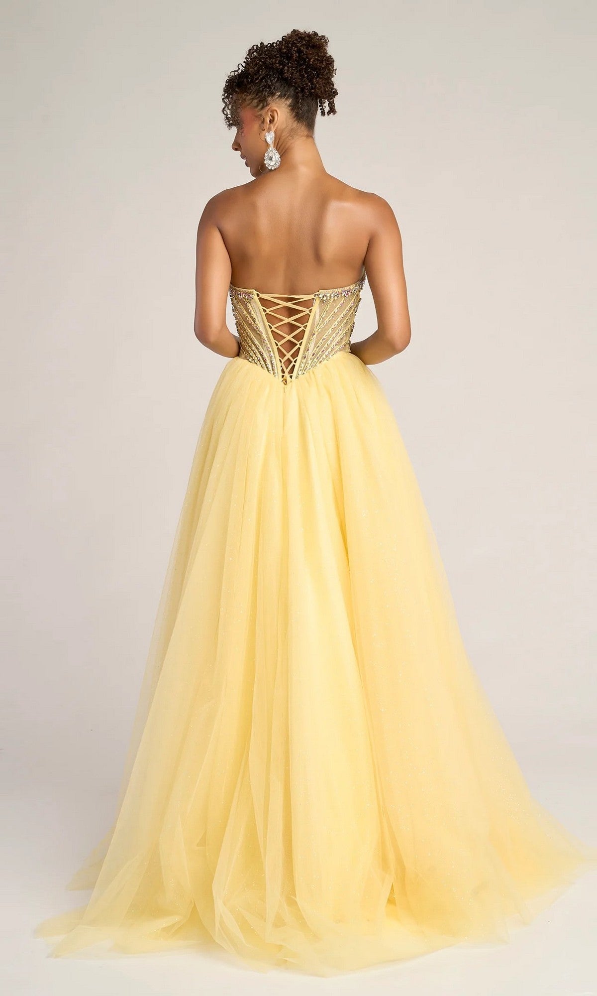 A woman stands facing away from the camera in the Portia and Scarlett XO047 long formal dress, featuring a strapless tulle ball gown skirt and a lace-up, beaded corset bodice—a stunning prom dress choice against a plain background.